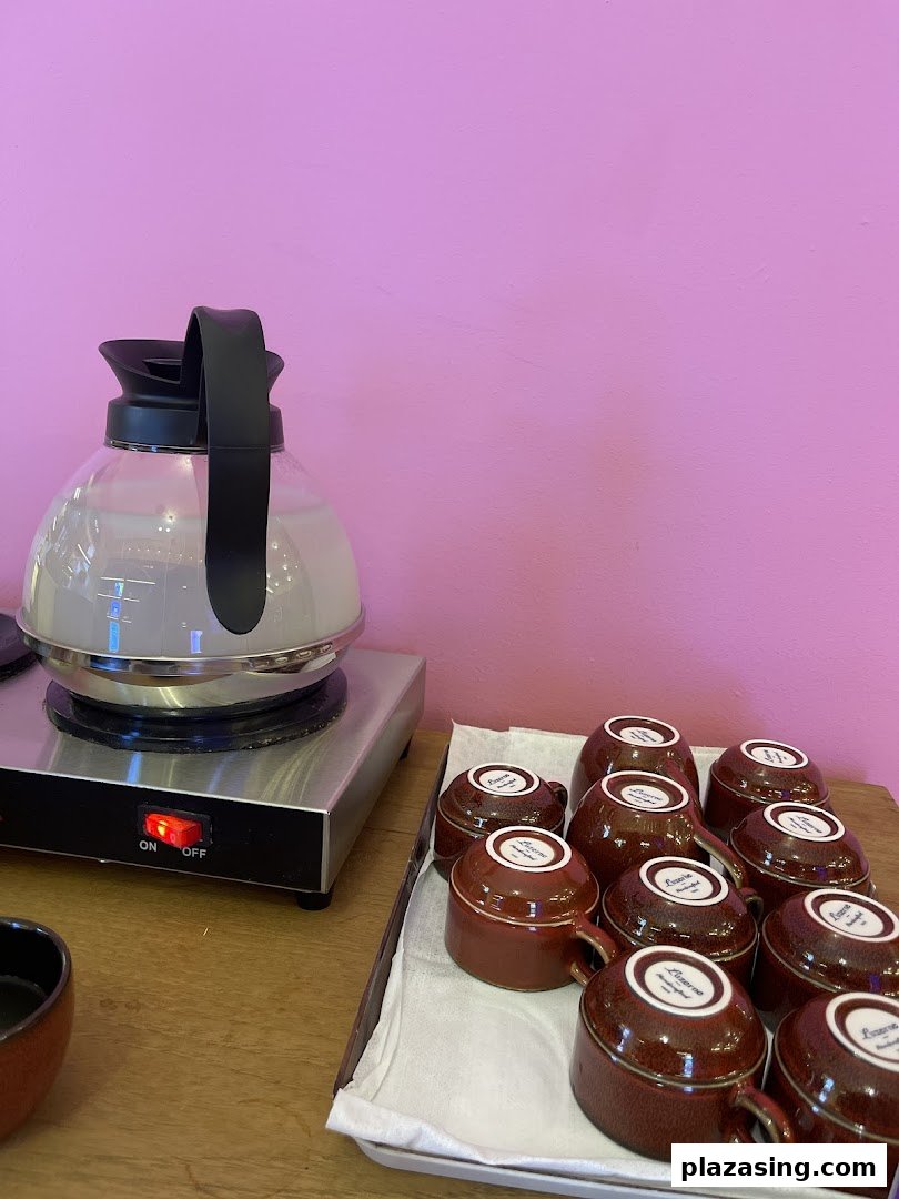 A kettle on a hot plate and a tray of ceramic cups against a vibrant pink wall, offering refreshments.