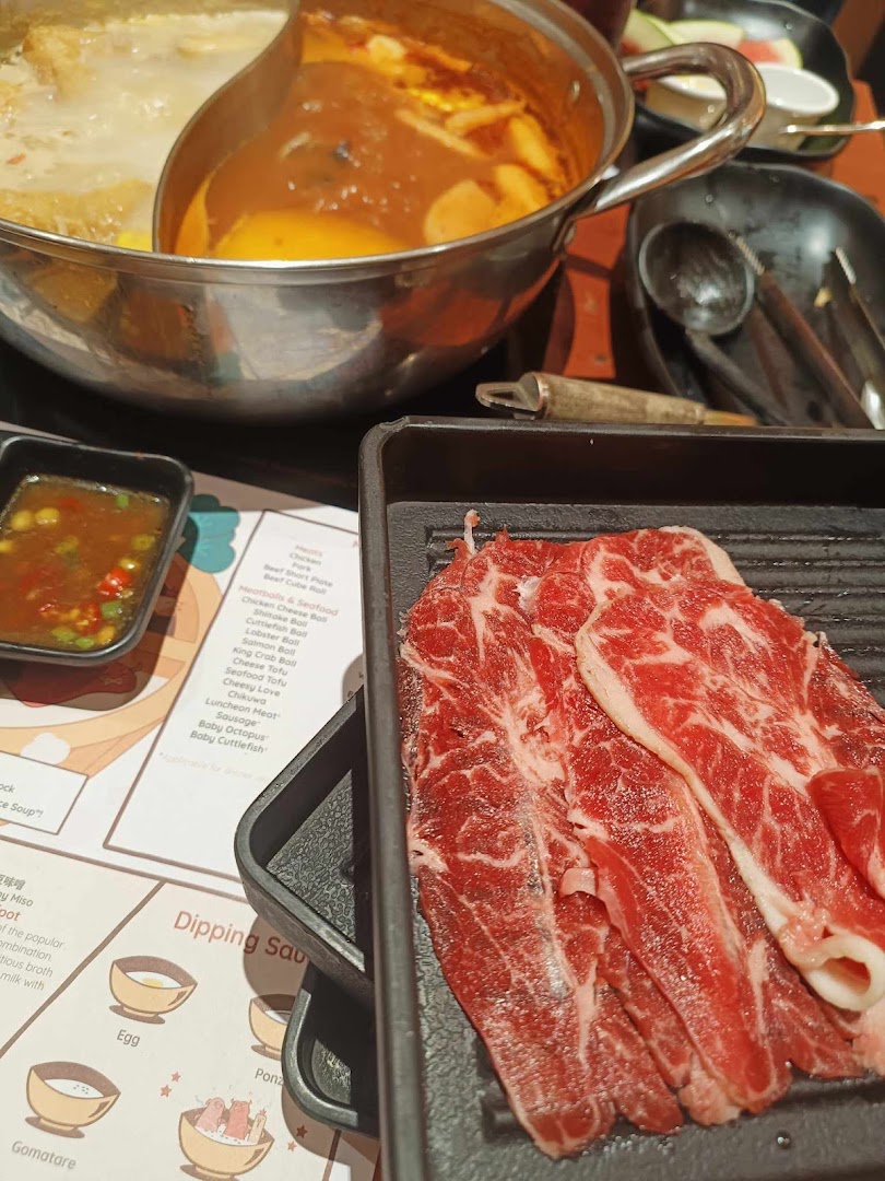 A close-up of a shabu-shabu hot pot with raw beef slices, dipping sauces, and a menu.