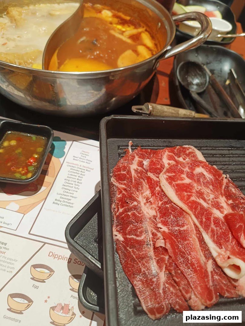 A close-up of a shabu-shabu hot pot with raw beef slices, dipping sauces, and a menu.
