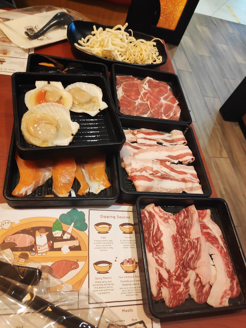 A table set with various fresh shabu-shabu ingredients including meats, seafood, noodles, and a placemat.