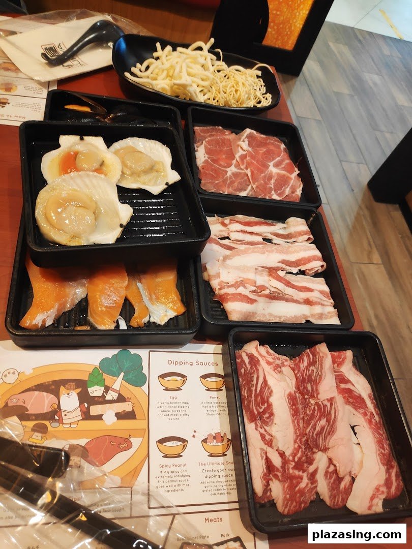A table set with various fresh shabu-shabu ingredients including meats, seafood, noodles, and a placemat.