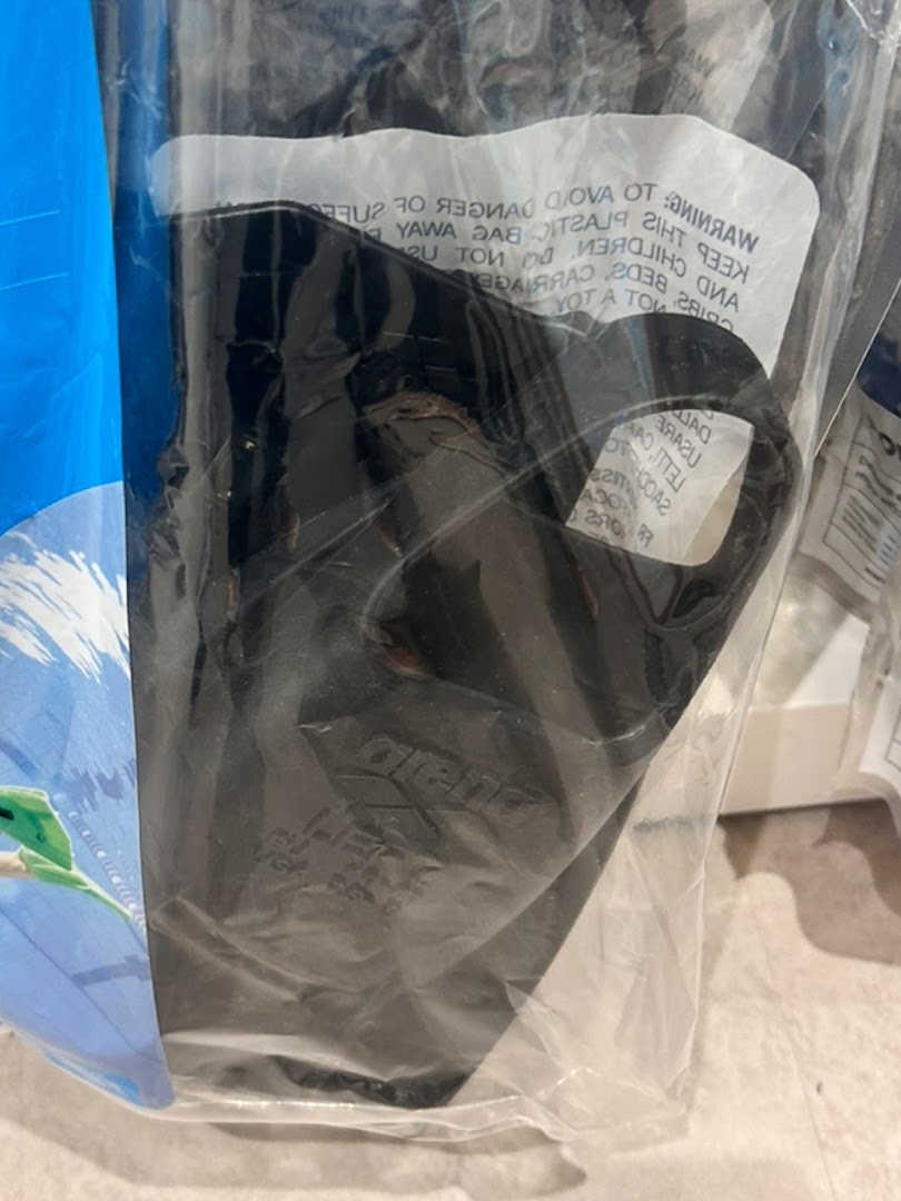 A black swimming fin or flipper is packaged in a clear plastic bag, with a warning label.