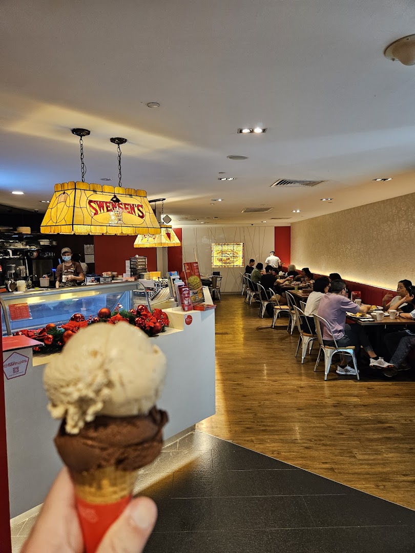 A hand holds a two-scoop ice cream cone inside Swensen's, with customers dining in the background.