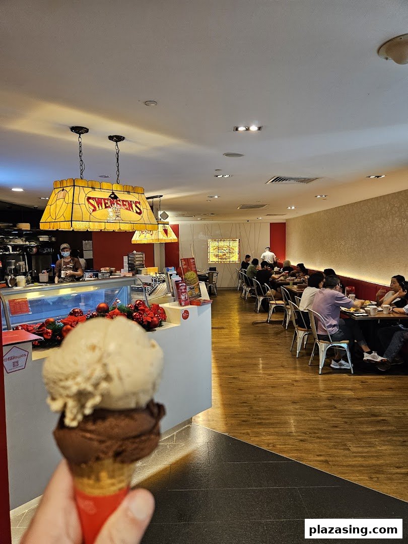 A hand holds a two-scoop ice cream cone inside Swensen's, with customers dining in the background.