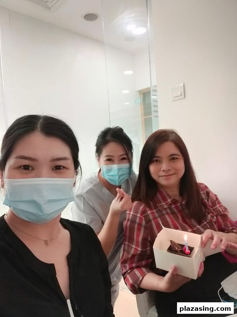 Three women, two masked, celebrate with a small cake and candle in a bright indoor setting.