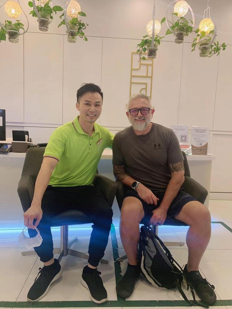 Two men smiling, seated in a bright, modern reception area with hanging plants.