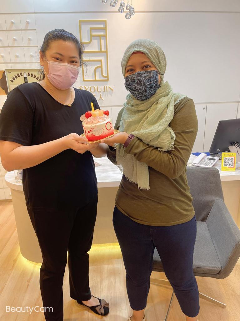 Two masked women, one staff and one customer, holding a small birthday cake inside Syoujin shop.
