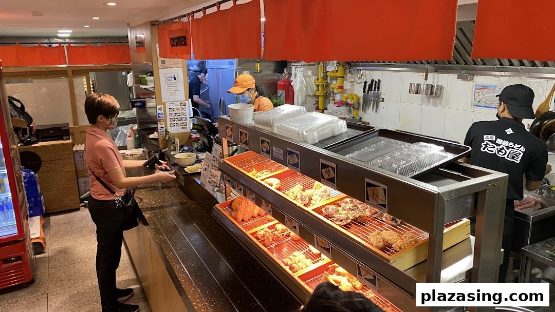 A customer orders at Tamoya Udon's counter, showcasing freshly prepared tempura and the bustling kitchen.