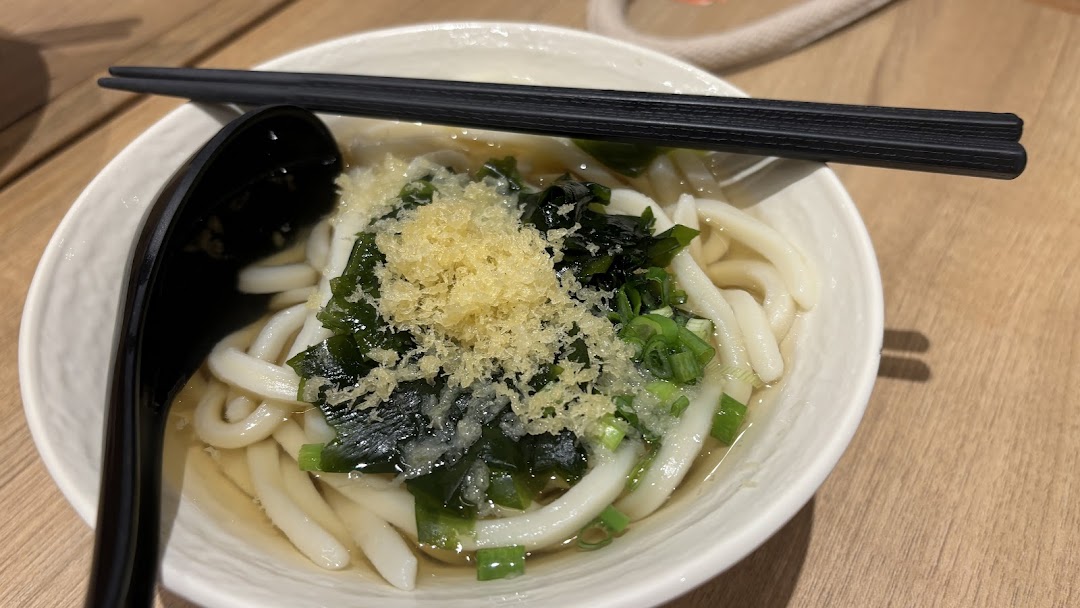 A close-up of a bowl of udon noodles with seaweed, green onions, and tempura flakes.