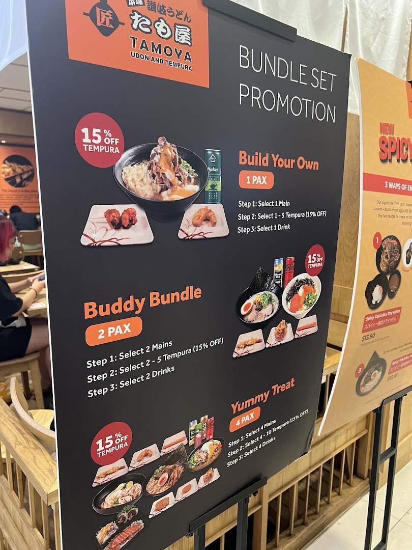 A promotional stand for Tamoya Udon & Tempura, showcasing various bundle set promotions and a new spicy udon dish.