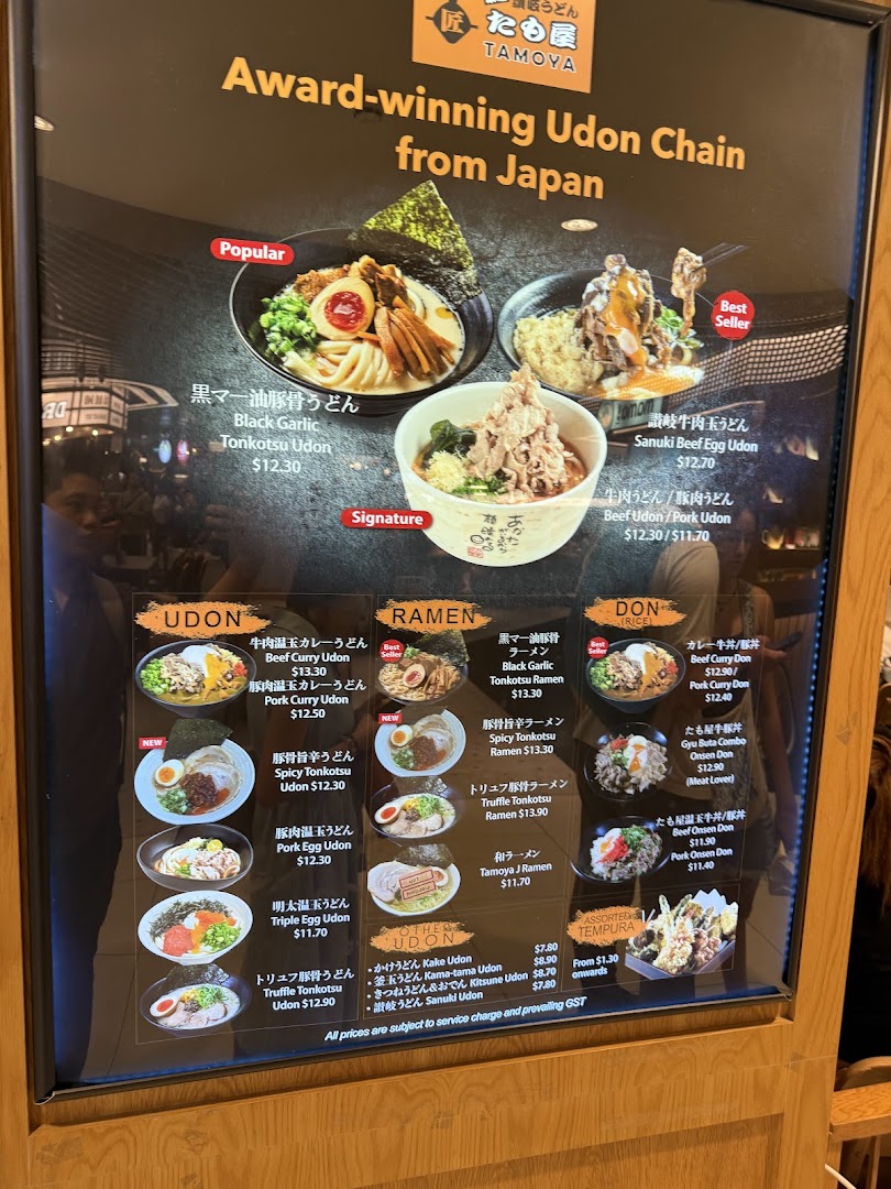 A brightly lit menu board showcasing a variety of udon, ramen, donburi, and tempura dishes with prices.