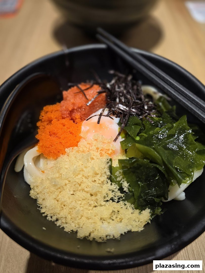 A vibrant bowl of udon noodles topped with tobiko, seaweed, tempura crumbs, and a poached egg.