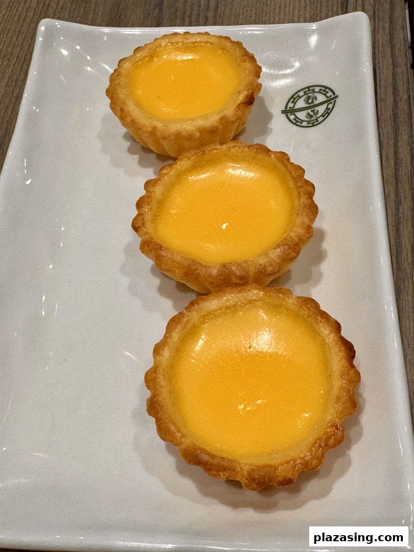 Three golden egg tarts are neatly arranged on a white rectangular plate, showcasing their delicious appearance.