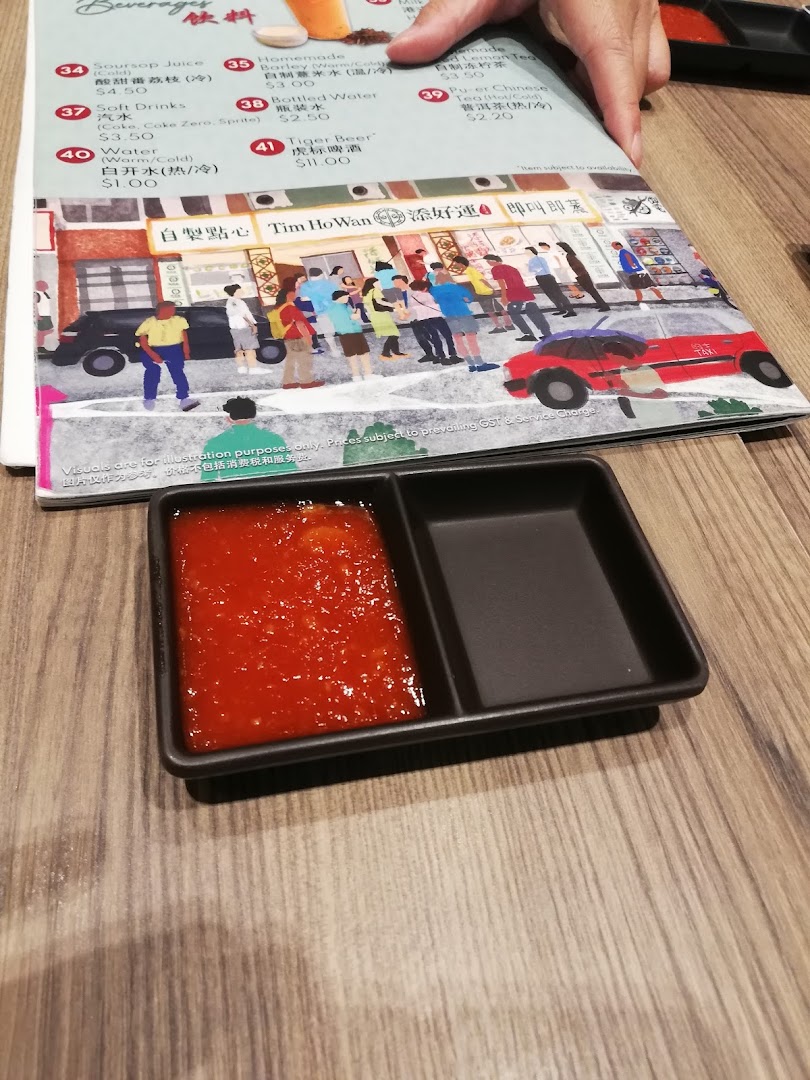 A Tim Ho Wan menu displaying beverages and prices, with a sauce dish on a wooden table.