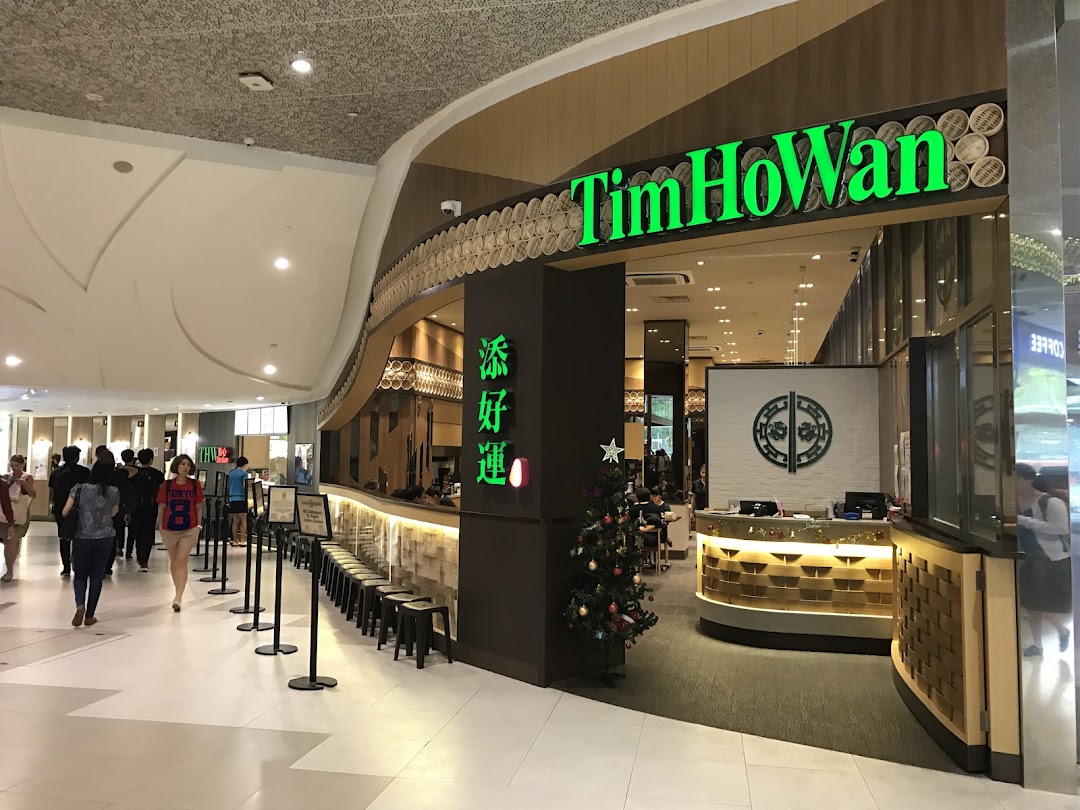 The vibrant green Tim Ho Wan shopfront welcomes diners in a bustling mall setting, featuring a festive Christmas tree.