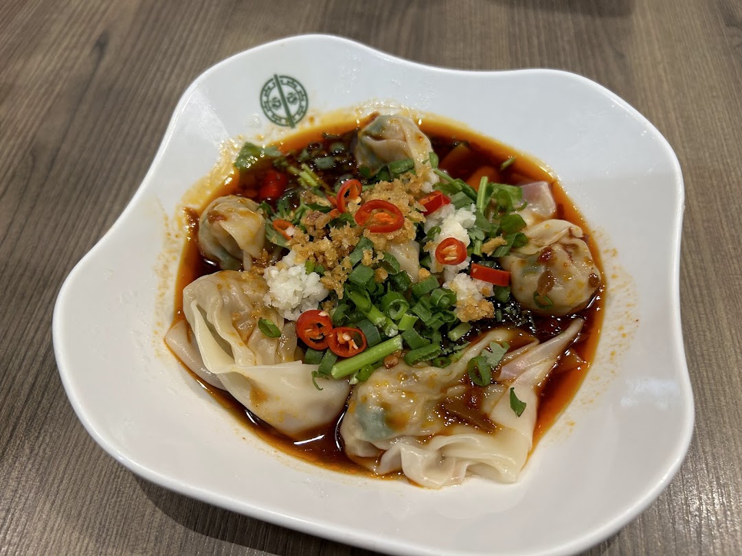 A close-up of spicy wontons or dumplings in a savory sauce, garnished with chili, garlic, and spring onions.