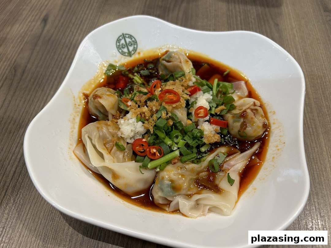 A close-up of spicy wontons or dumplings in a savory sauce, garnished with chili, garlic, and spring onions.