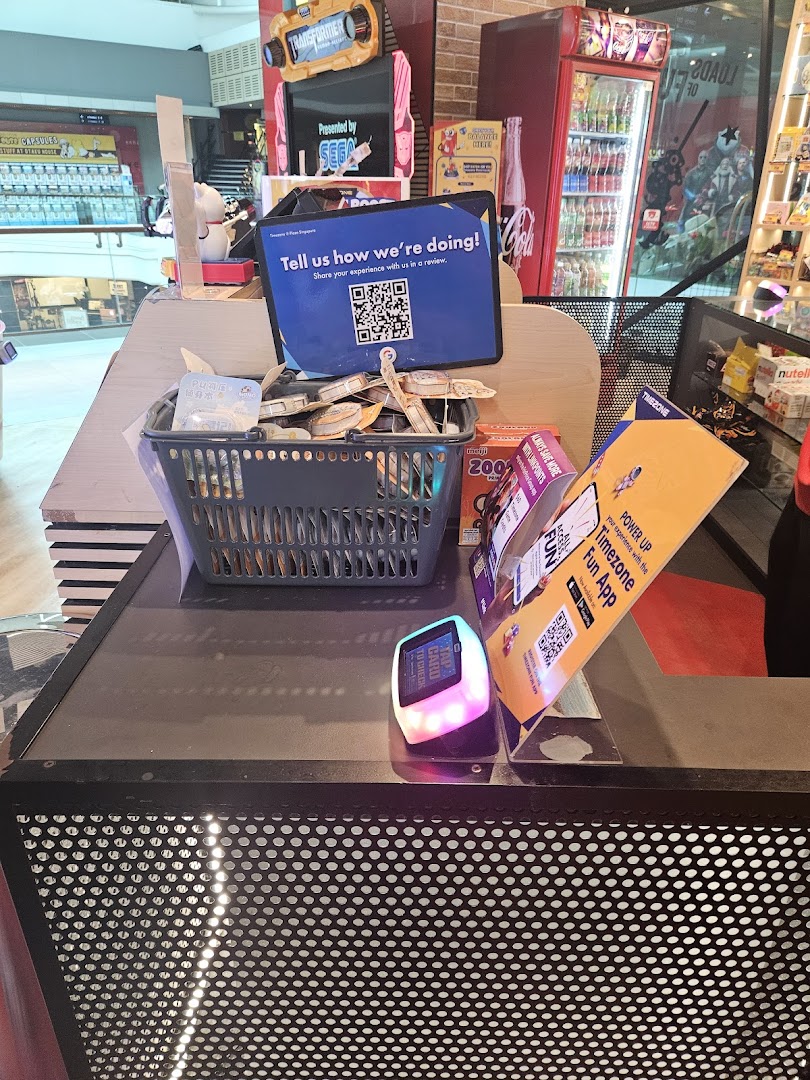 A counter at Timezone arcade with a customer feedback sign, prize basket, and arcade games in the background.
