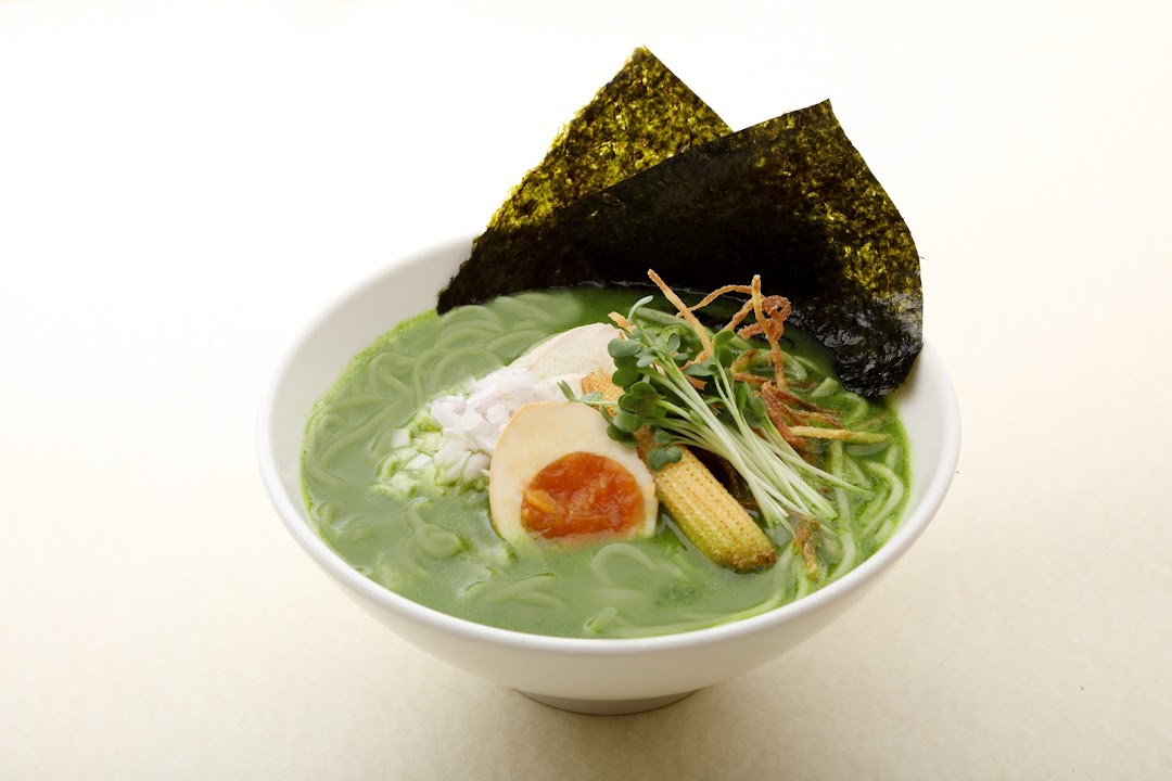 A vibrant green ramen bowl featuring noodles, egg, baby corn, sprouts, and nori seaweed.