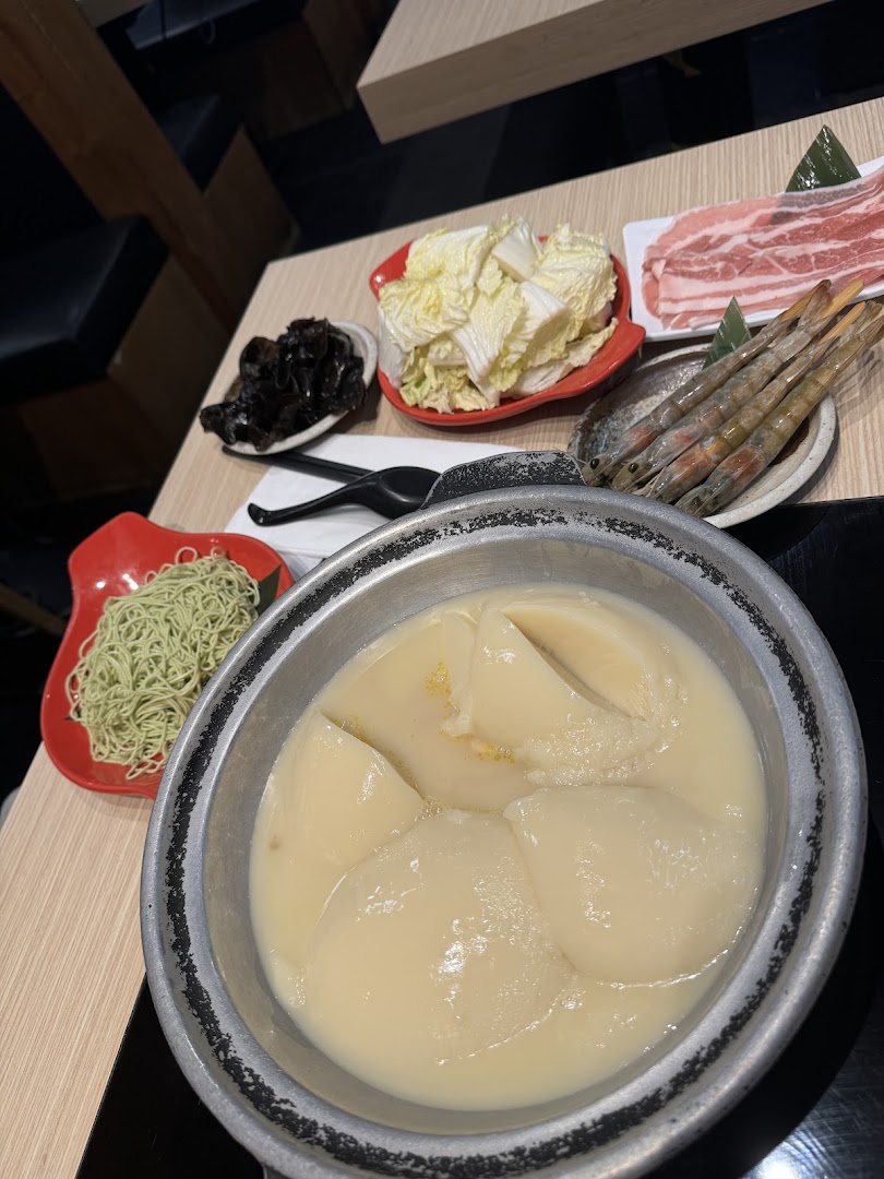 A hotpot setup featuring a collagen-rich chicken broth, fresh vegetables, raw meat, shrimp, and green noodles.