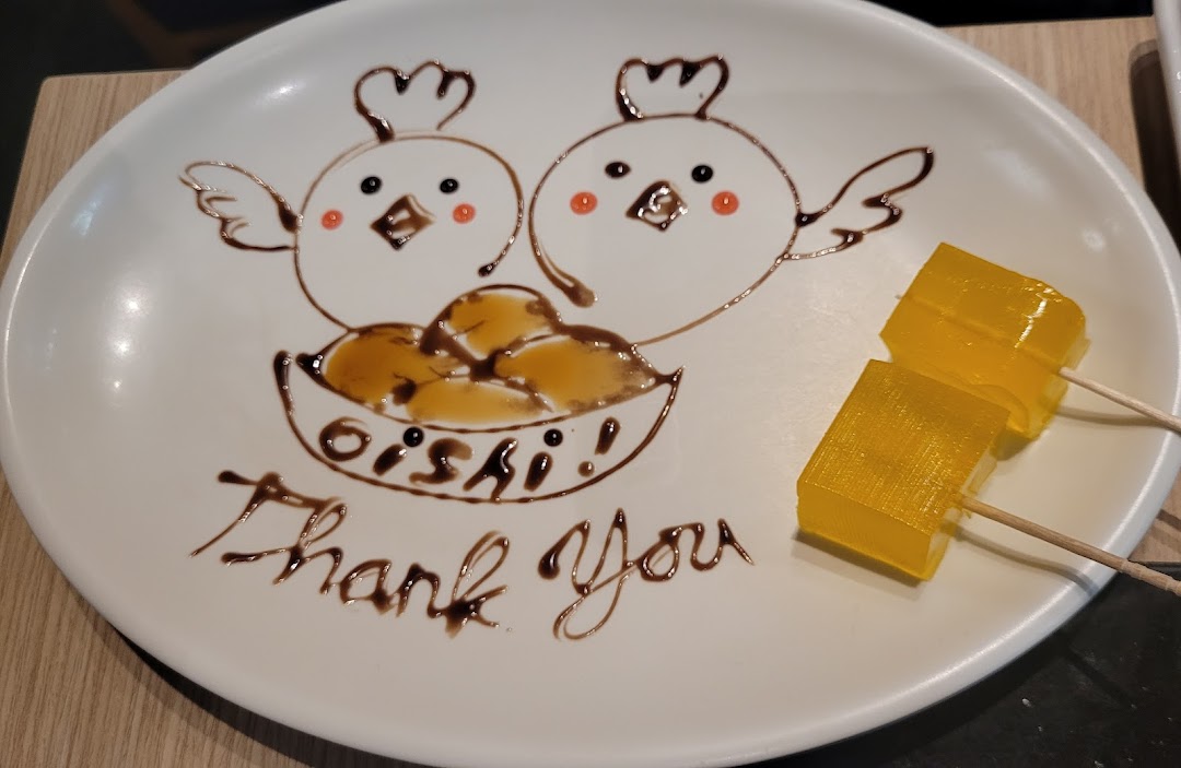A white dessert plate features chocolate art of two chicks, "OISHI!", "Thank You", and two yellow jelly cubes.