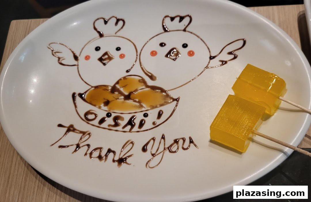 A white dessert plate features chocolate art of two chicks, "OISHI!", "Thank You", and two yellow jelly cubes.