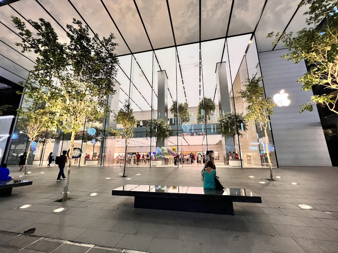 Modern glass-fronted Apple Store with trees and people outside, illuminated at dusk.