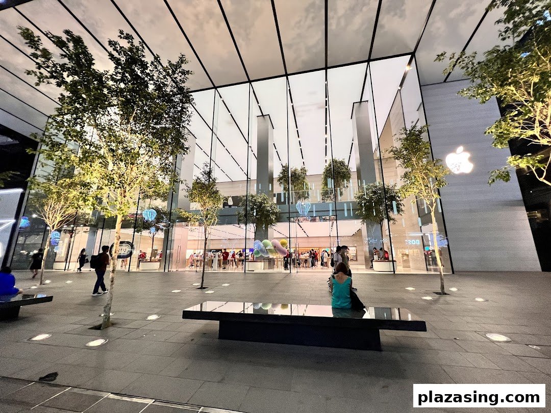 Modern glass-fronted Apple Store with trees and people outside, illuminated at dusk.