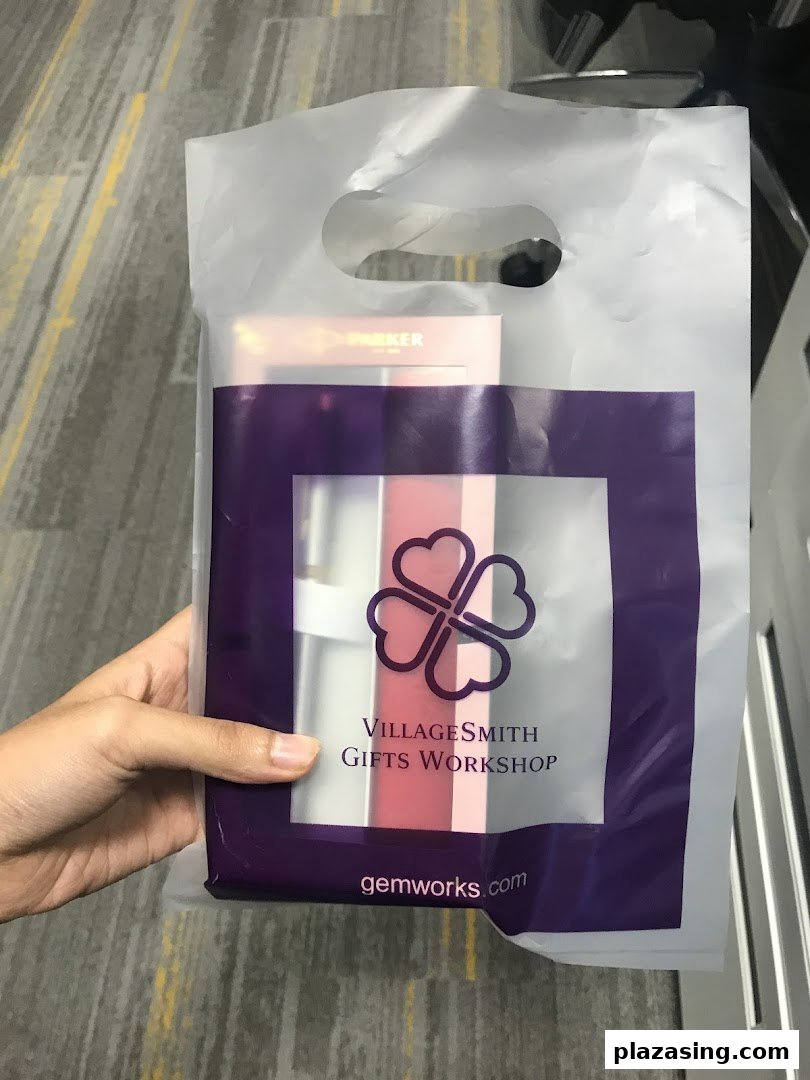 A hand holds a transparent shopping bag from VillageSmith Gifts Workshop, containing a gift item.