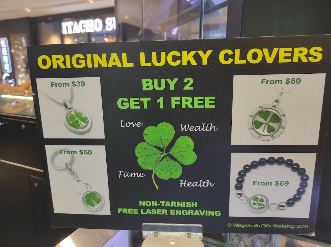 A promotional sign for VillageSmith Gifts Workshop, showcasing 'Original Lucky Clovers' jewelry and accessories with prices and a 'Buy 2 Get 1 Free' offer.
