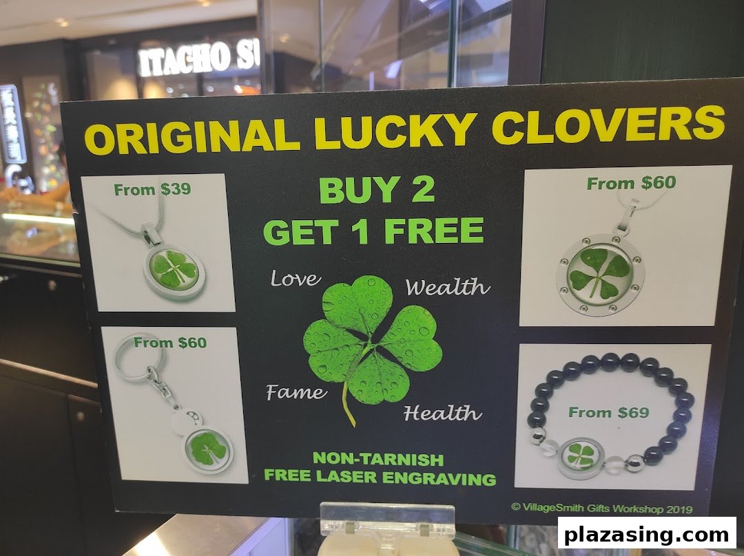 A promotional sign for VillageSmith Gifts Workshop, showcasing 'Original Lucky Clovers' jewelry and accessories with prices and a 'Buy 2 Get 1 Free' offer.