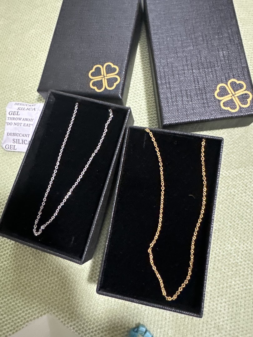 Two delicate necklaces, one silver and one gold, displayed in black gift boxes with a clover logo.