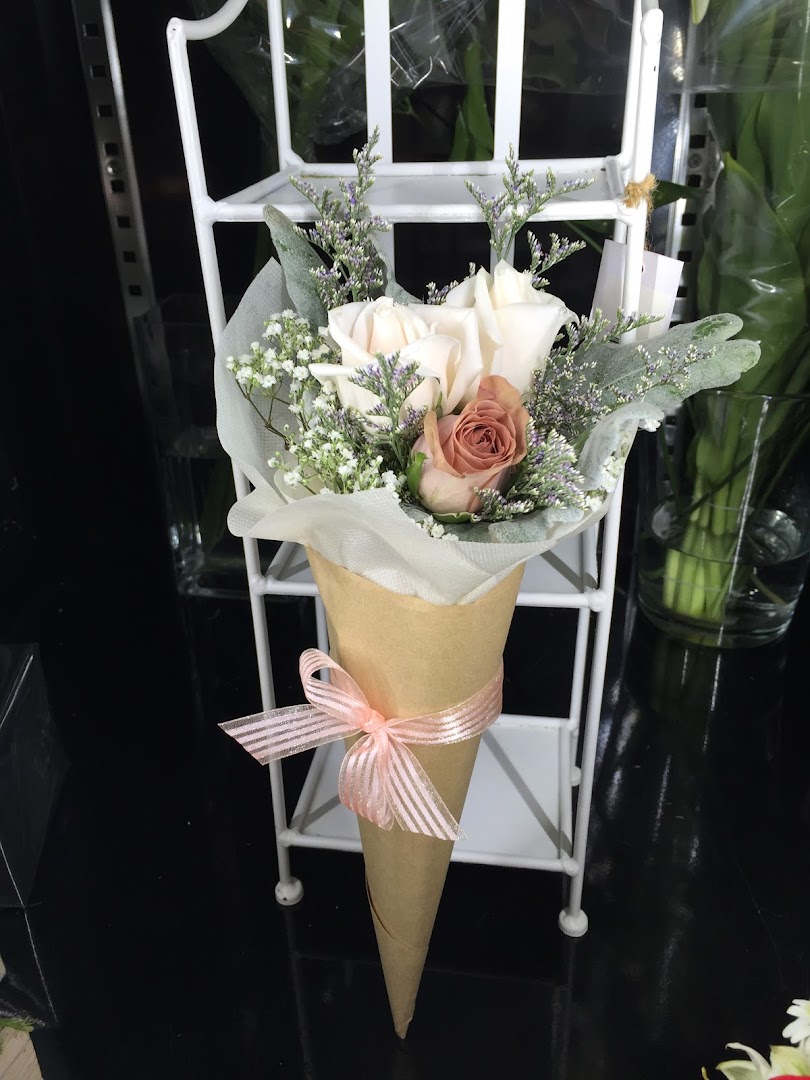 A charming bouquet of roses and foliage in a paper cone with a pink ribbon, displayed on a white stand.