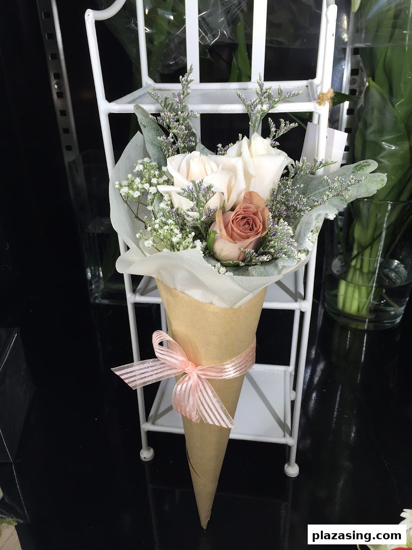 A charming bouquet of roses and foliage in a paper cone with a pink ribbon, displayed on a white stand.