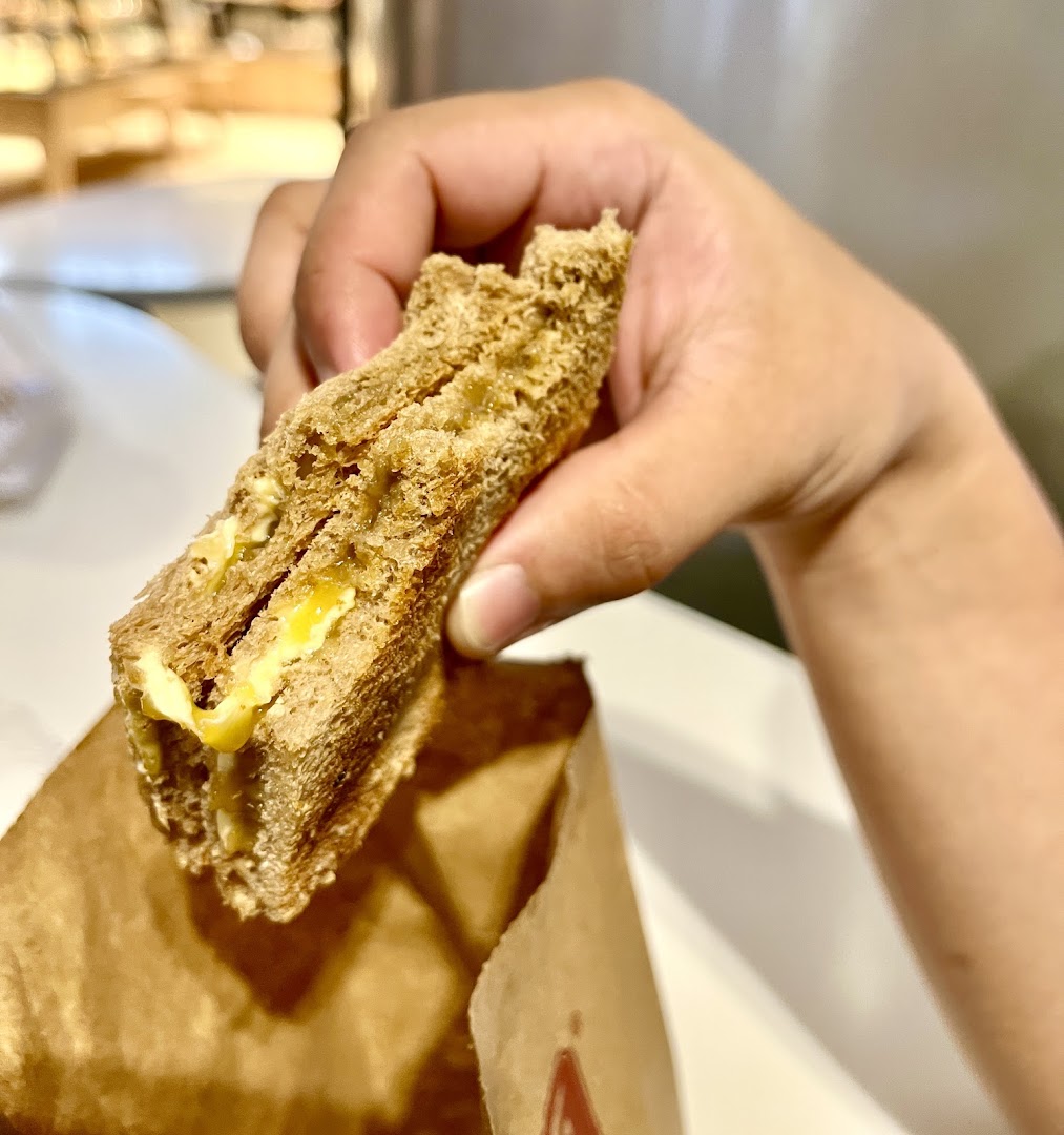 A hand holds a piece of toasted kaya toast with butter, emerging from a brown paper bag.