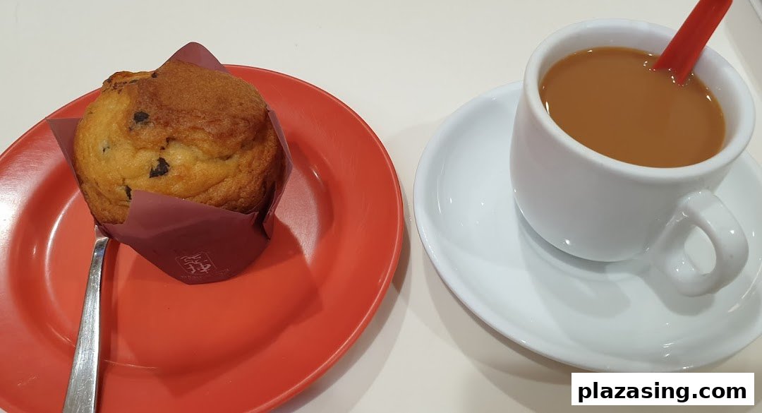 A delicious chocolate chip muffin on a red plate next to a cup of coffee or tea.