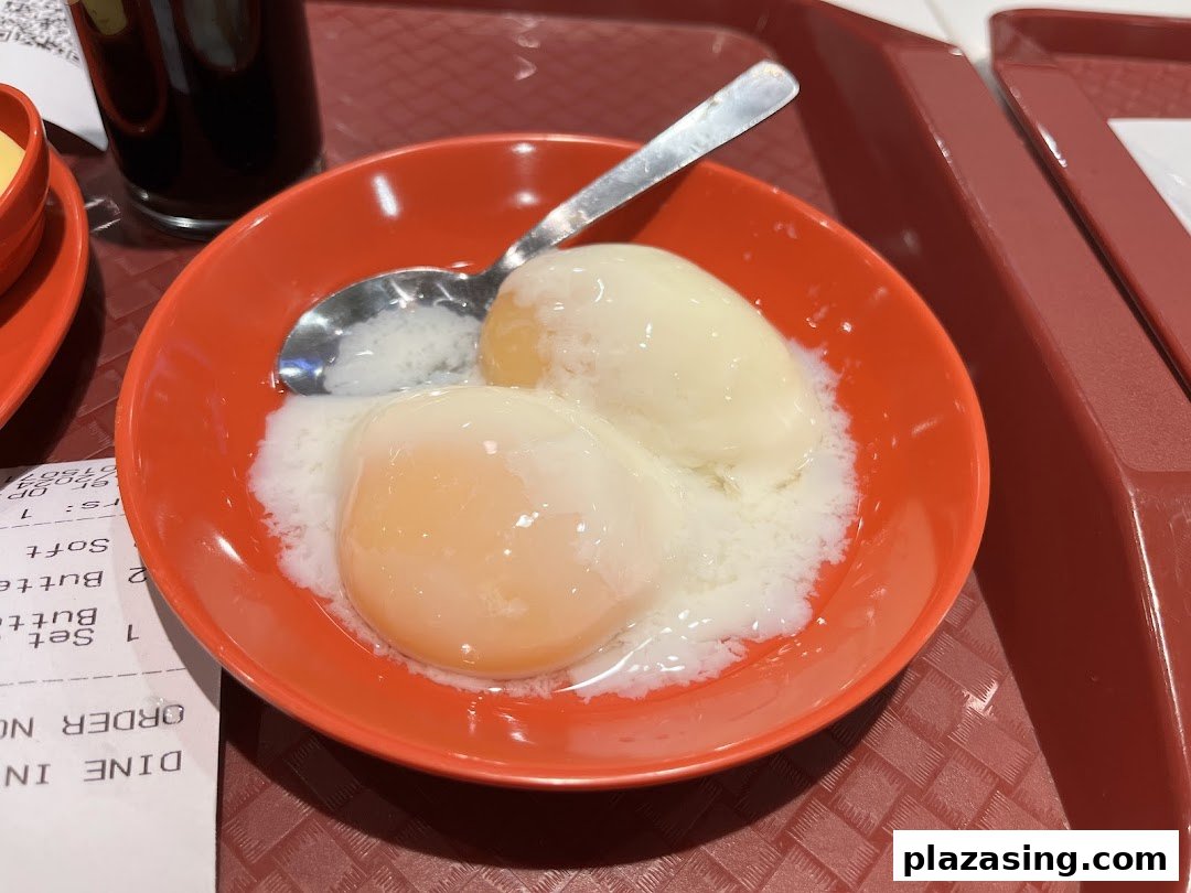 A close-up of traditional soft-boiled eggs in an orange bowl with a spoon, served on a red tray.