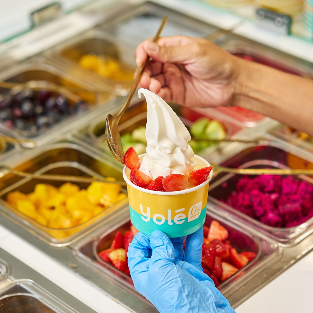 A hand in a blue glove holds a Yolé frozen yogurt cup with strawberries, surrounded by fresh fruit toppings.