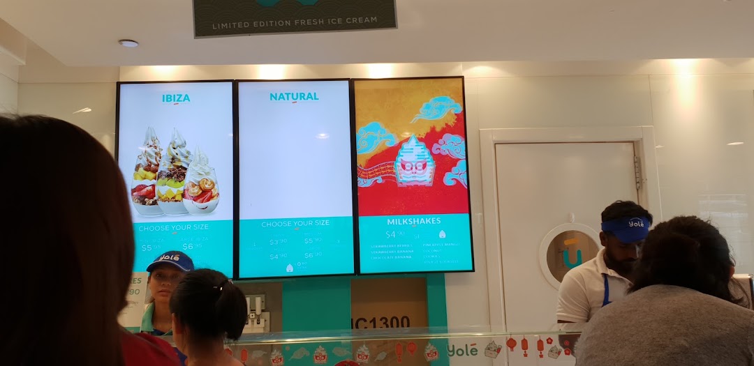 Digital menu boards at Yolé displaying frozen yogurt and milkshake options with prices and customers at the counter.