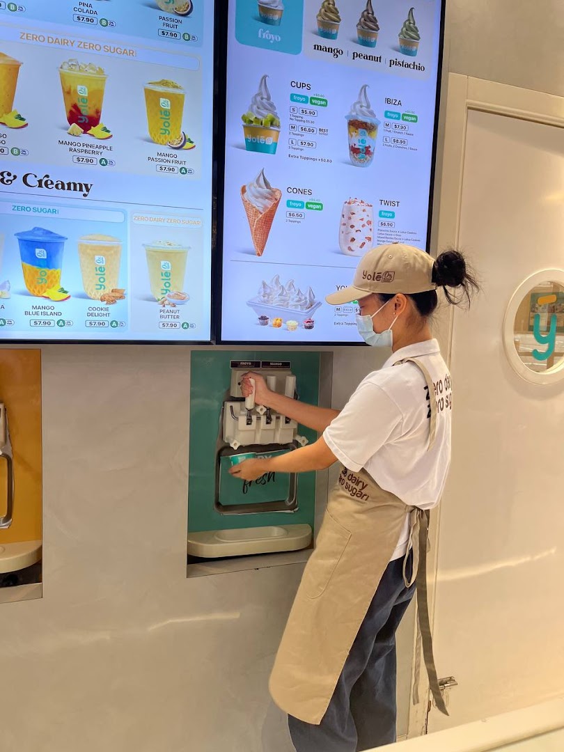 A staff member prepares a frozen dessert at Yolé, with digital menu boards displaying various frozen yogurt and drink options.