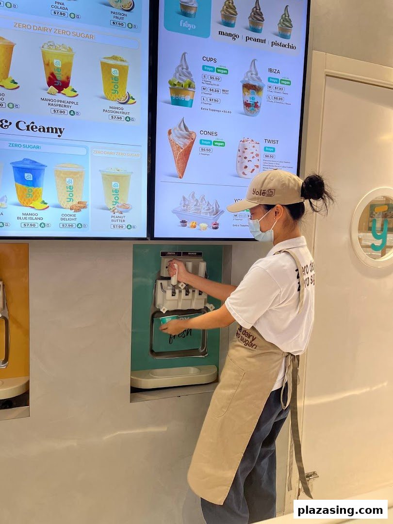 A staff member prepares a frozen dessert at Yolé, with digital menu boards displaying various frozen yogurt and drink options.