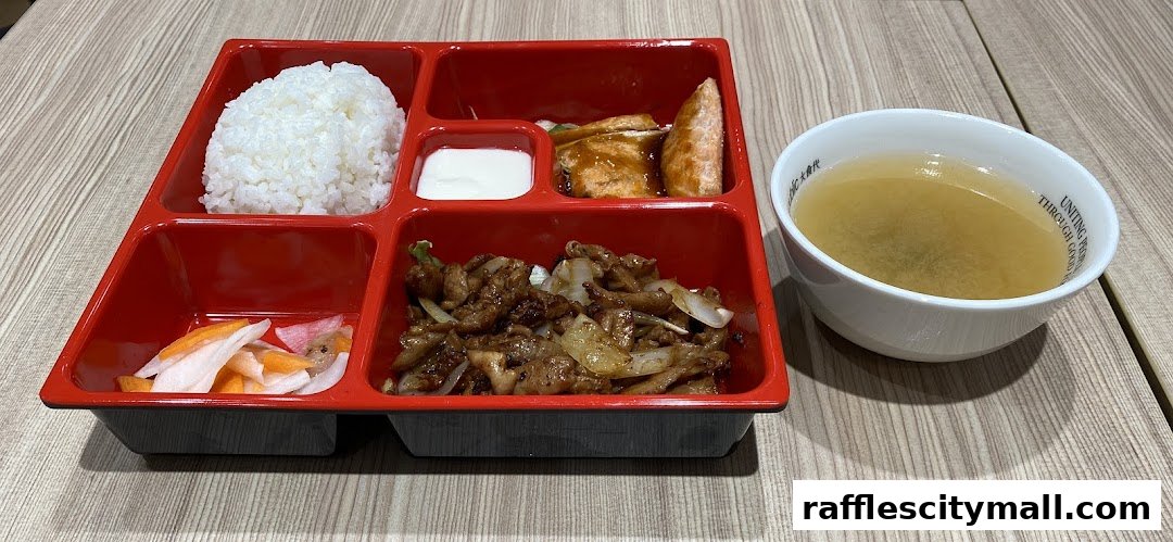 A bento box meal with rice, stir-fried meat, pickled vegetables, and a side of soup.