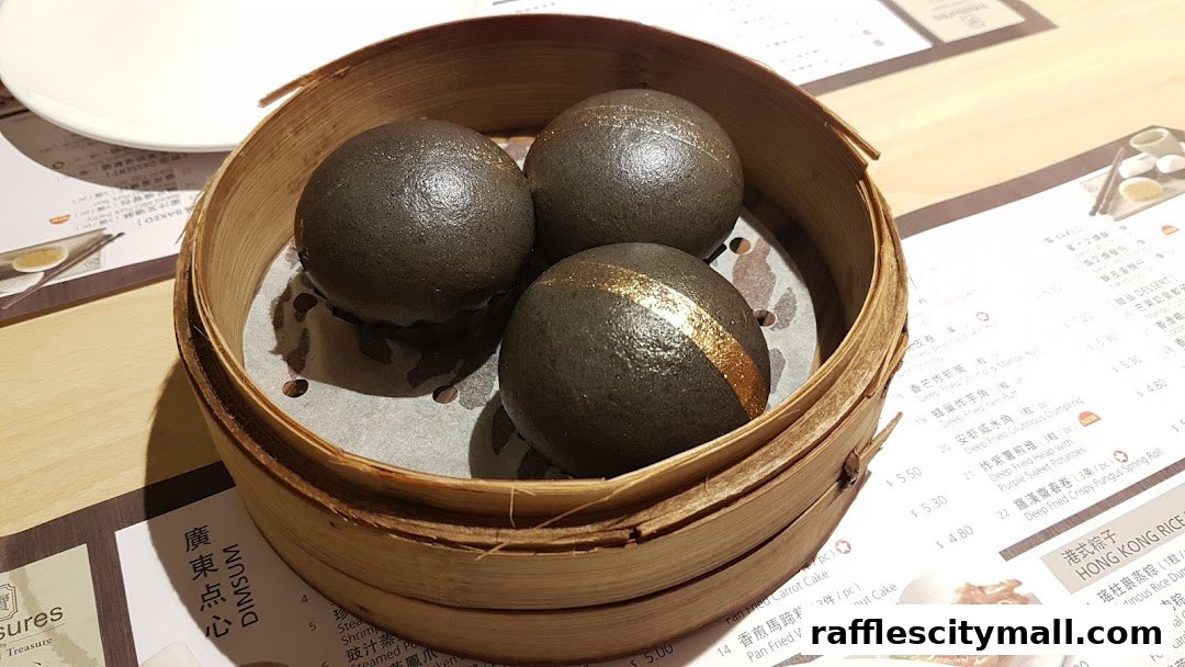 A bamboo steamer basket filled with three dark, round dim sum buns, one with a gold stripe.