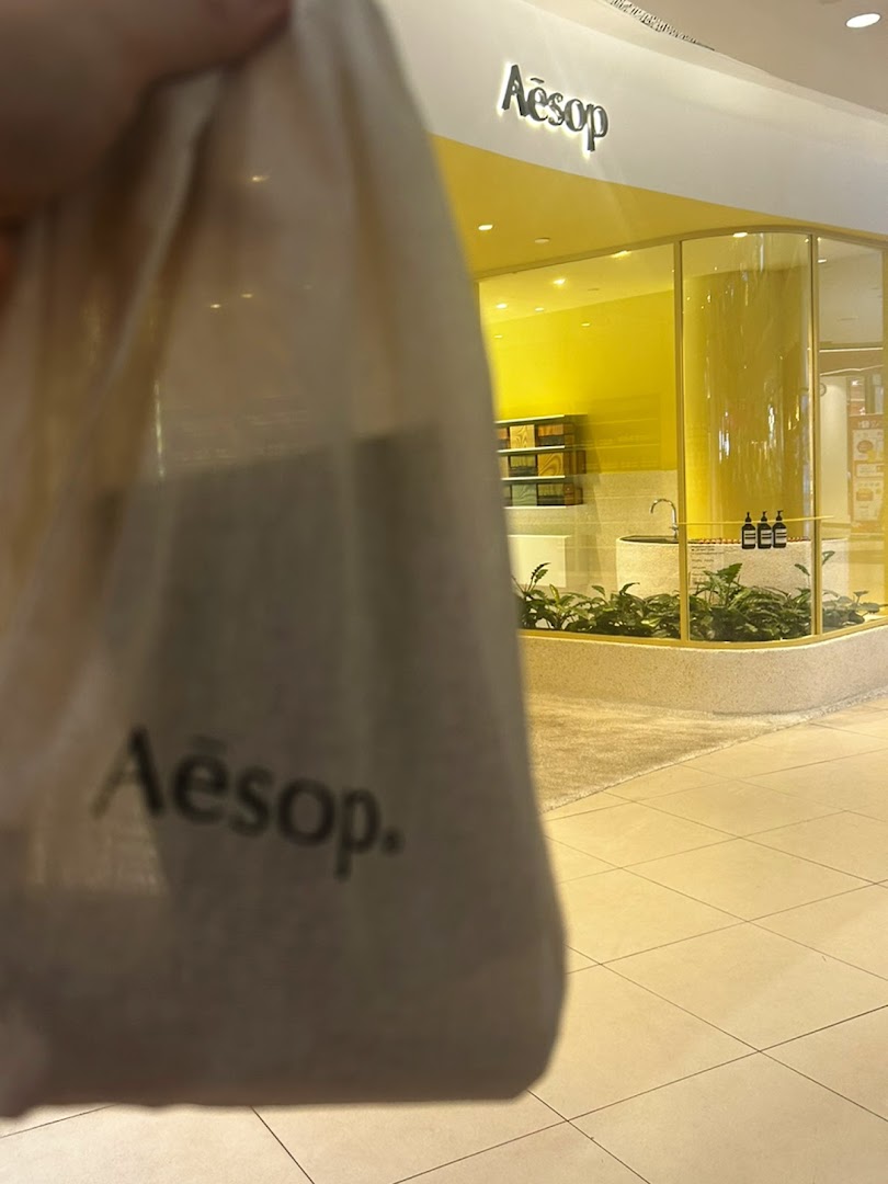 A person holds an Aesop bag in front of the bright yellow Aesop store entrance.