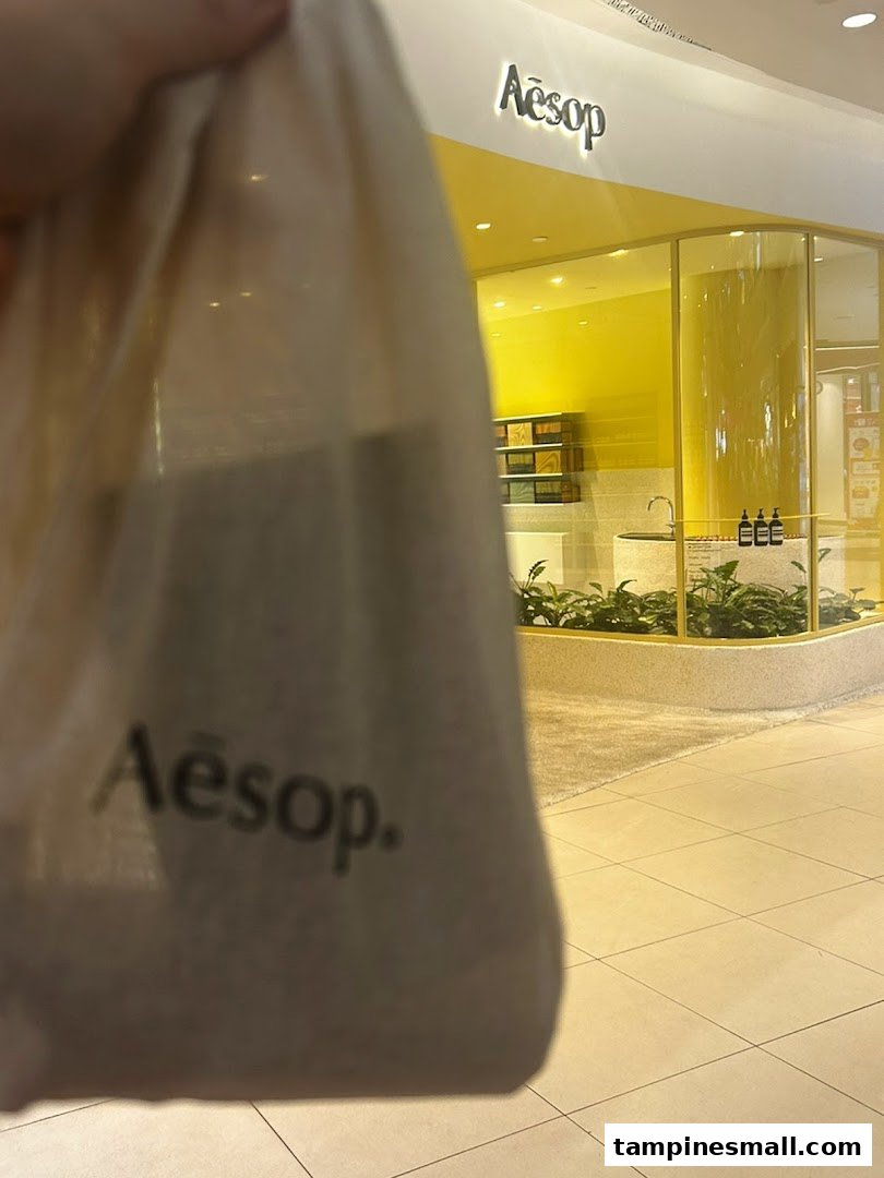 A person holds an Aesop bag in front of the bright yellow Aesop store entrance.