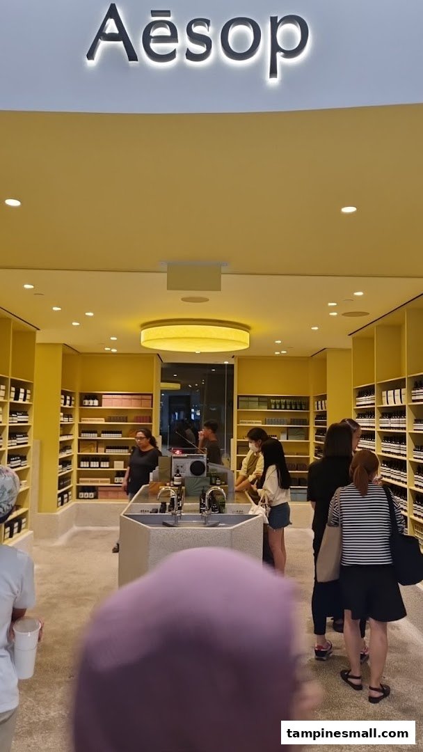 The interior of an Aesop store featuring its distinctive product displays, a central sink area, and customers browsing.