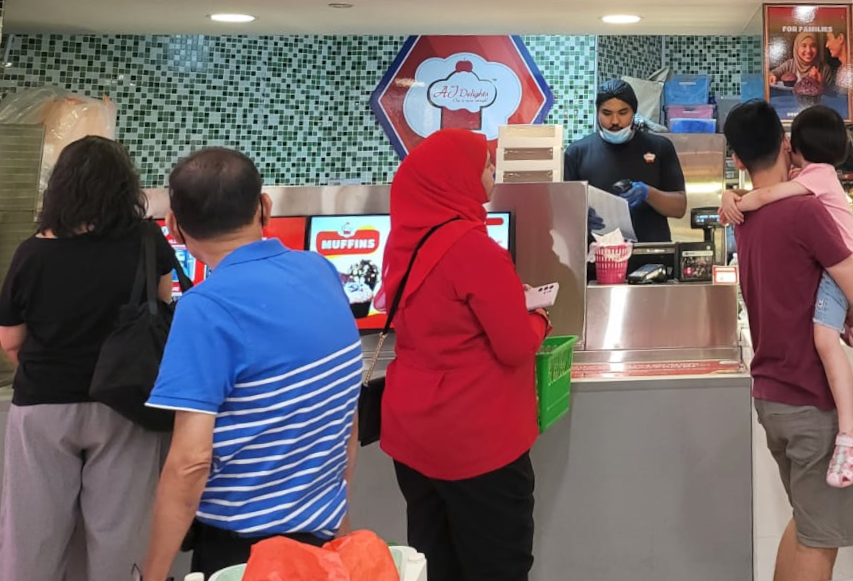 Customers queue at the counter of AJ Delights, served by a staff member in a bustling shop interior.