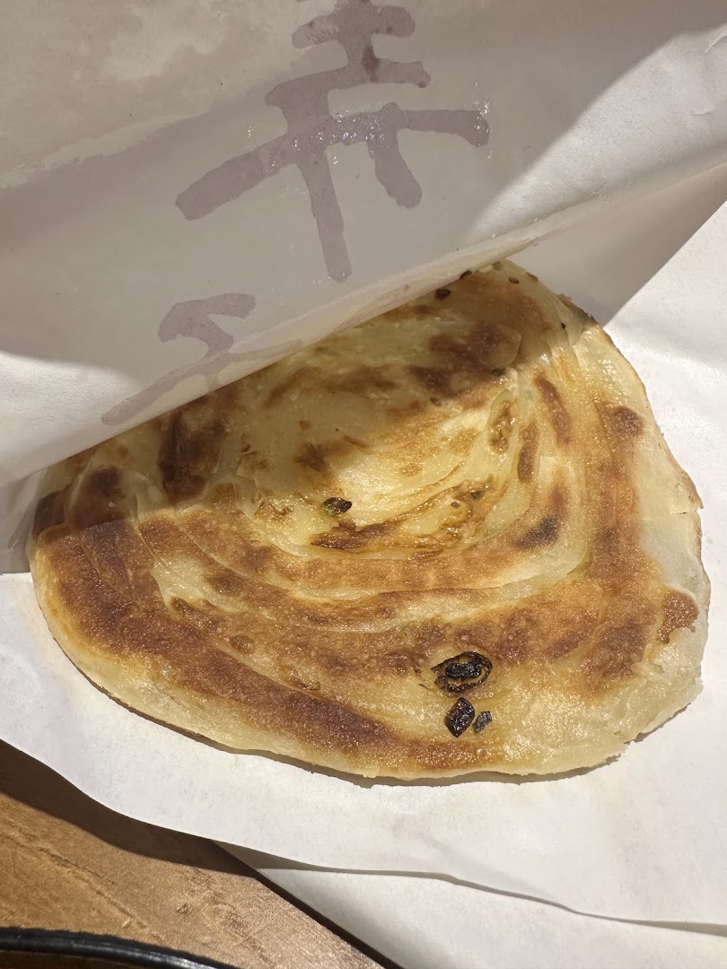 A golden-brown, flaky scallion pancake served in a paper wrapper from Alley Wei.