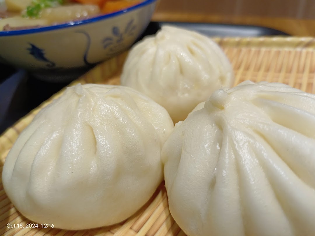 Close-up of three fluffy steamed buns on a bamboo tray, with a bowl of stew in the background.