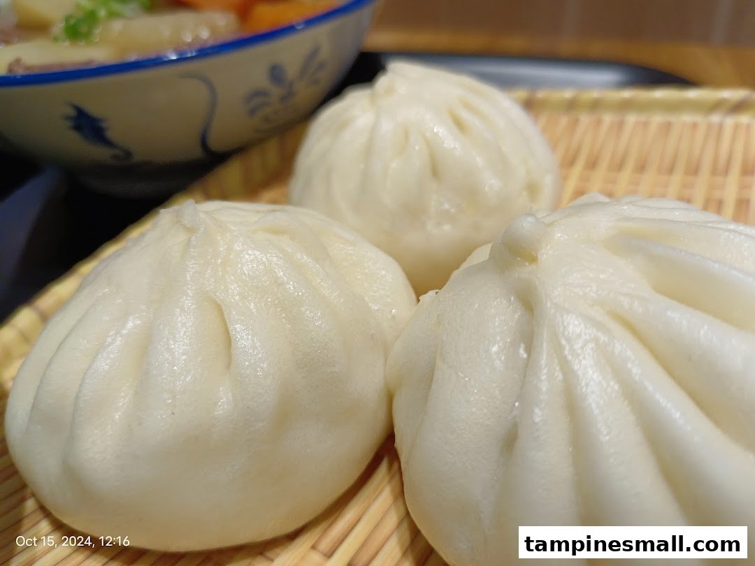 Close-up of three fluffy steamed buns on a bamboo tray, with a bowl of stew in the background.